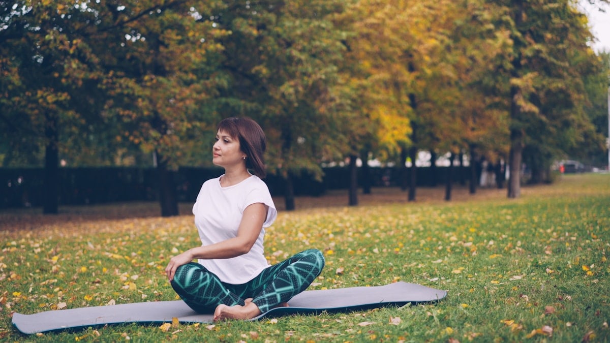 Flexible girl yogini is practising simple twist pose then resting in lotus position sitting on yoga mat on grass in city park. Recreational area and people concept.