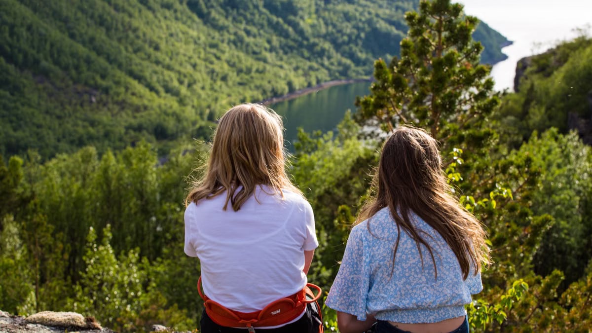 Two women sit overlooking a lush mountain landscape in Troms og Finnmark, Norway, on a sunny day.